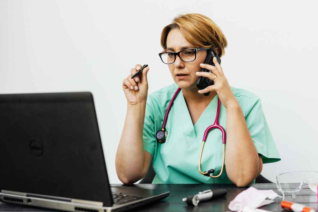 Woman doctor sitting at desk on the phone and looking at a laptop